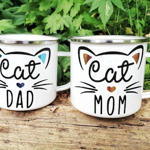 May include: Two white enamel mugs with black cat faces and the words "Cat Dad" and "Cat Mom" in black lettering. The mugs have silver rims.