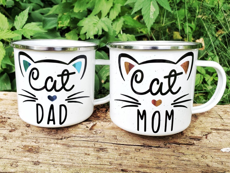 May include: Two white enamel mugs with black cat faces and the words "Cat Dad" and "Cat Mom" in black lettering. The mugs have silver rims.