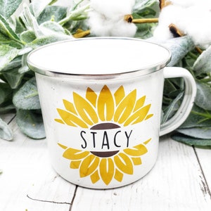 Op de afbeelding: Witte emaille mok met een geel zonnebloemontwerp en de naam "Stacy" in zwart op een bruin lint.
