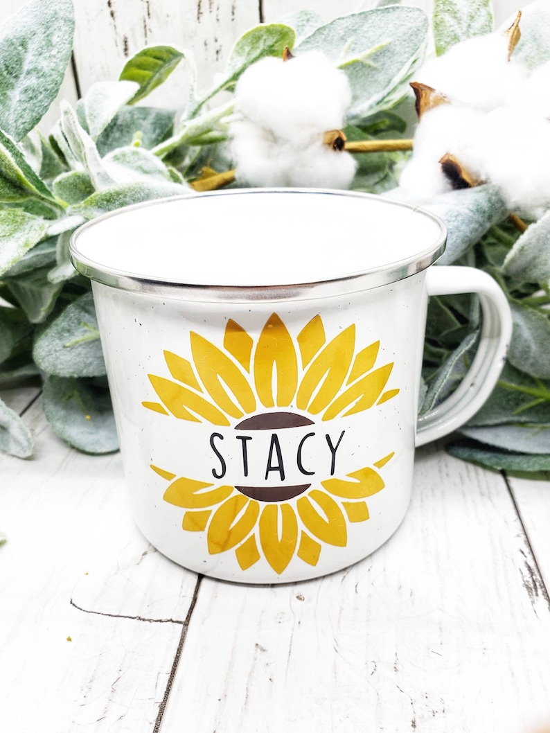 Op de afbeelding: Witte emaille mok met een geel zonnebloemontwerp en de naam "Stacy" in zwart op een bruin lint.