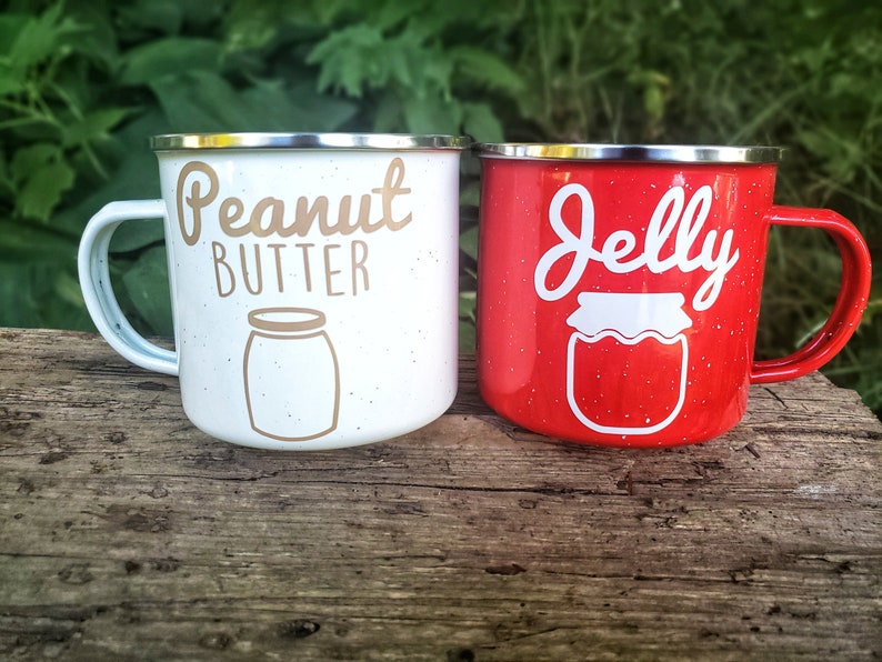 May include: Two enamel mugs with a speckled finish. One mug is white with the text "Peanut Butter" and a brown outline of a jar. The other mug is red with the text "Jelly" and a white outline of a jar.