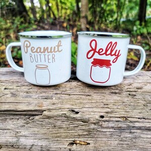 May include: Two white enamel mugs with black rims. The mug on the left says "Peanut Butter" in brown lettering with a white outline and a drawing of a peanut butter jar. The mug on the right says "Jelly" in red lettering with a white outline and a drawing of a jelly jar.