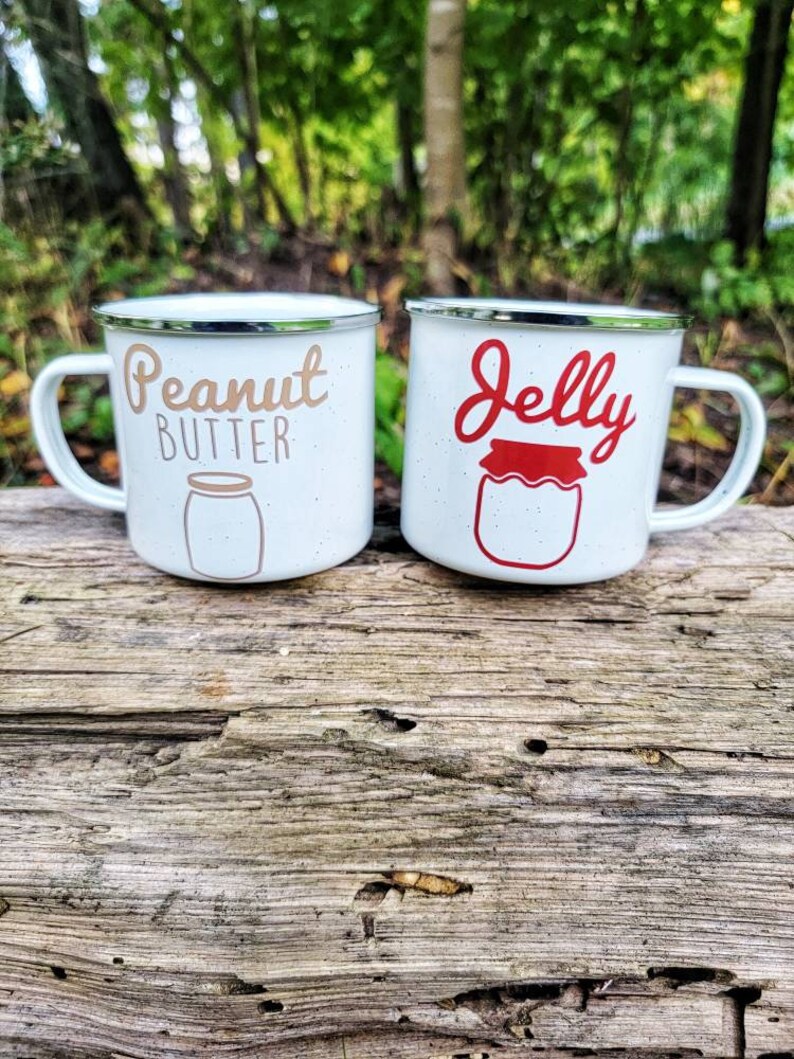 May include: Two white enamel mugs with black rims. The mug on the left says "Peanut Butter" in brown lettering with a white outline and a drawing of a peanut butter jar. The mug on the right says "Jelly" in red lettering with a white outline and a drawing of a jelly jar.