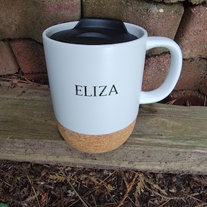 May include: White travel mug with a black lid and a cork base. The name "ELIZA" is printed in black on the front of the mug. The mug has a white handle and is sitting on a wooden surface.