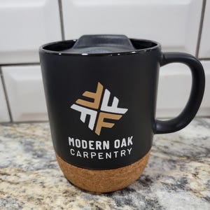 May include: Black travel mug with a cork bottom and a white logo that says "Modern Oak Carpentry" with a geometric design in gold.
