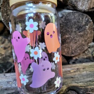 May include: A clear glass reusable water bottle with a wooden lid and a straw. The bottle is decorated with pink, orange, and white ghost and flower stickers.