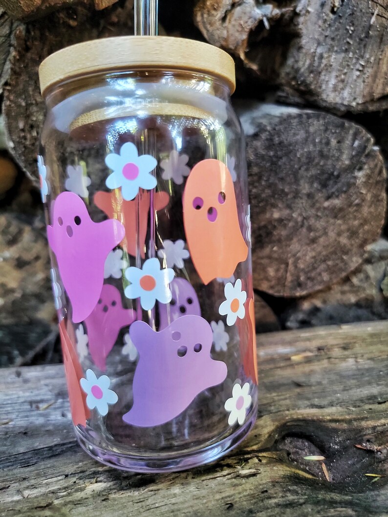 May include: A clear glass reusable water bottle with a wooden lid and a straw. The bottle is decorated with pink, orange, and white ghost and flower stickers.