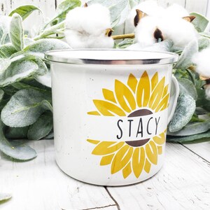 Op de afbeelding: Witte emaille mok met een geel zonnebloemontwerp en de naam "Stacy" op een bruin lint.