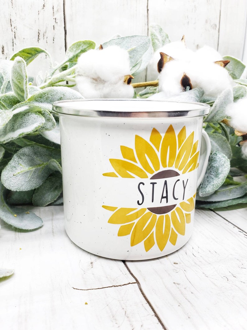 Op de afbeelding: Witte emaille mok met een geel zonnebloemontwerp en de naam "Stacy" op een bruin lint.
