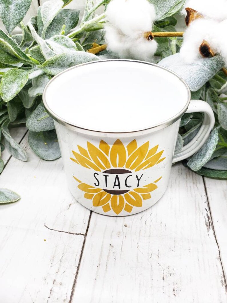Op de afbeelding: Witte emaille mok met een zilveren rand en een geel zonnebloemontwerp. De zonnebloem heeft een bruin centrum met de naam "Stacy" in zwart.