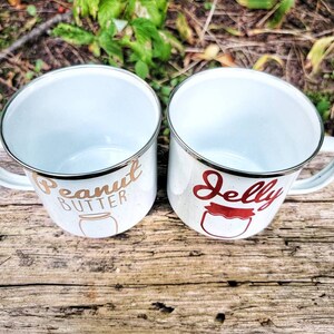 May include: Two white enamel mugs with gold and red lettering. The mug on the left says "Peanut Butter" with a gold jar illustration. The mug on the right says "Jelly" with a red jar illustration.