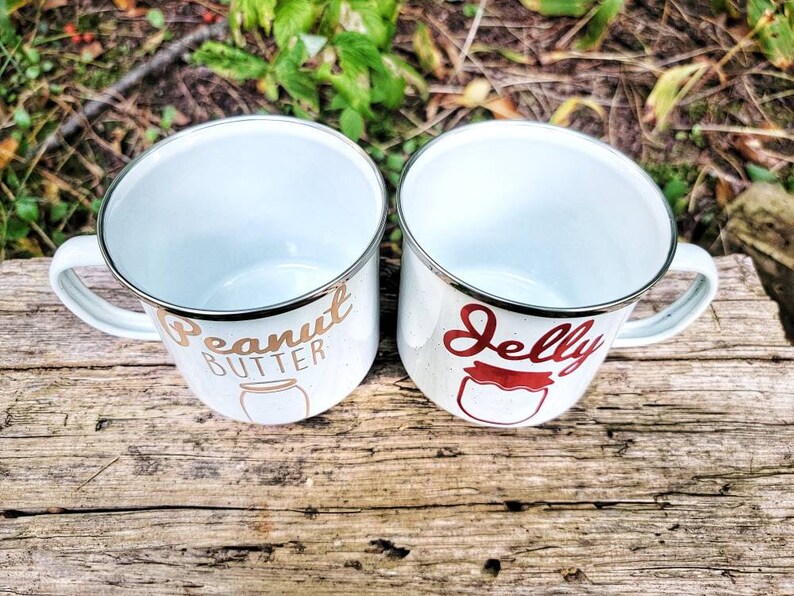 May include: Two white enamel mugs with gold and red lettering. The mug on the left says "Peanut Butter" with a gold jar illustration. The mug on the right says "Jelly" with a red jar illustration.