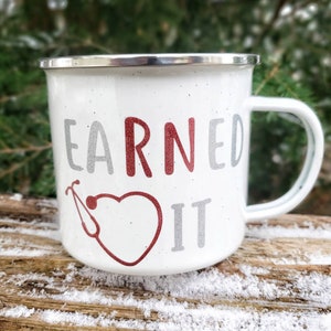 May include: White enamel mug with a red heart and a stethoscope design. The text "EARNED IT" is written in gray and red.