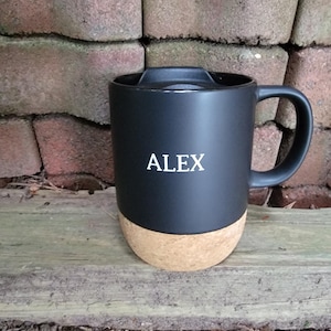 May include: Black travel mug with a cork base and a black handle. The word "ALEX" is printed in white on the front of the mug. The mug is sitting on a wooden surface in front of a brick wall.
