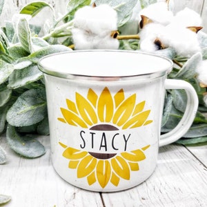 Op de afbeelding: Witte emaille campingmok met een geel zonnebloemontwerp en de naam "Stacy" gedrukt op de voorkant.