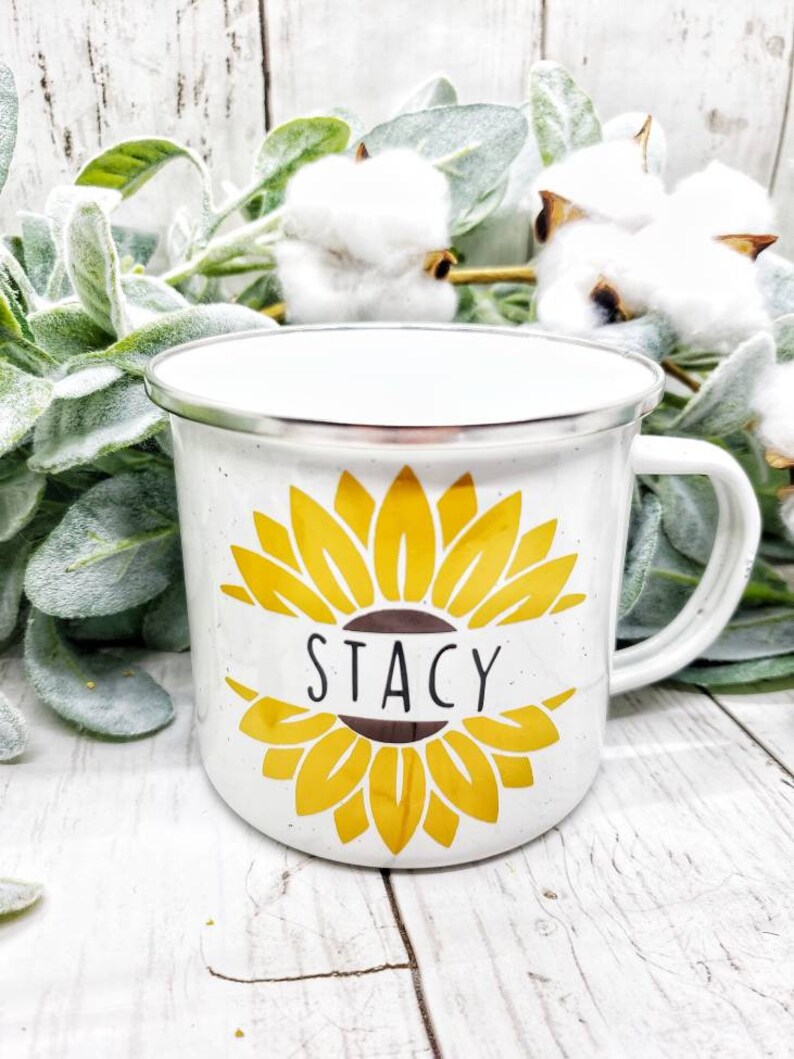 Op de afbeelding: Witte emaille campingmok met een geel zonnebloemontwerp en de naam "Stacy" gedrukt op de voorkant.