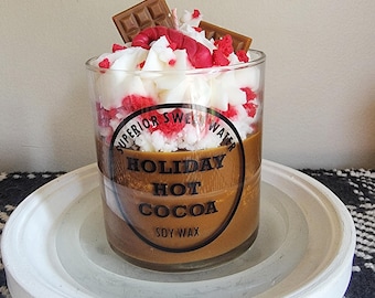 May include: A soy wax candle in a clear glass, resembling a hot cocoa drink. The candle has a brown base, topped with white and red wax, and chocolate bar accents. The glass is on a white, round tray.