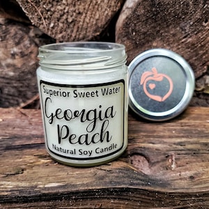 May include: A clear glass jar containing a natural soy candle with a white label that reads Superior Sweet Water Georgia Peach Natural Soy Candle. The silver metal lid features a peach graphic with a small heart inside the fruit outline.