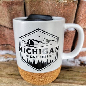 May include: White ceramic travel mug with a cork bottom. The mug features a black and white graphic of a mountain range, a sailboat, and the text "Michigan - Est. 1837 - "