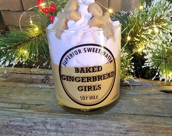 May include: A clear glass candle with a light brown wax base and a pink top layer, topped with gingerbread men. The candle has a label that reads "Baked Gingerbread Girls Soy Wax". The candle is surrounded by festive greenery and lights.