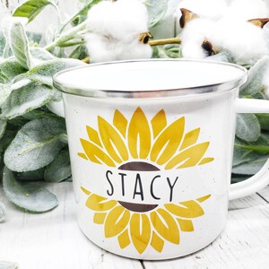 Op de afbeelding: Witte emaille mok met een geel zonnebloemontwerp en de naam "Stacy" op een bruin lint.