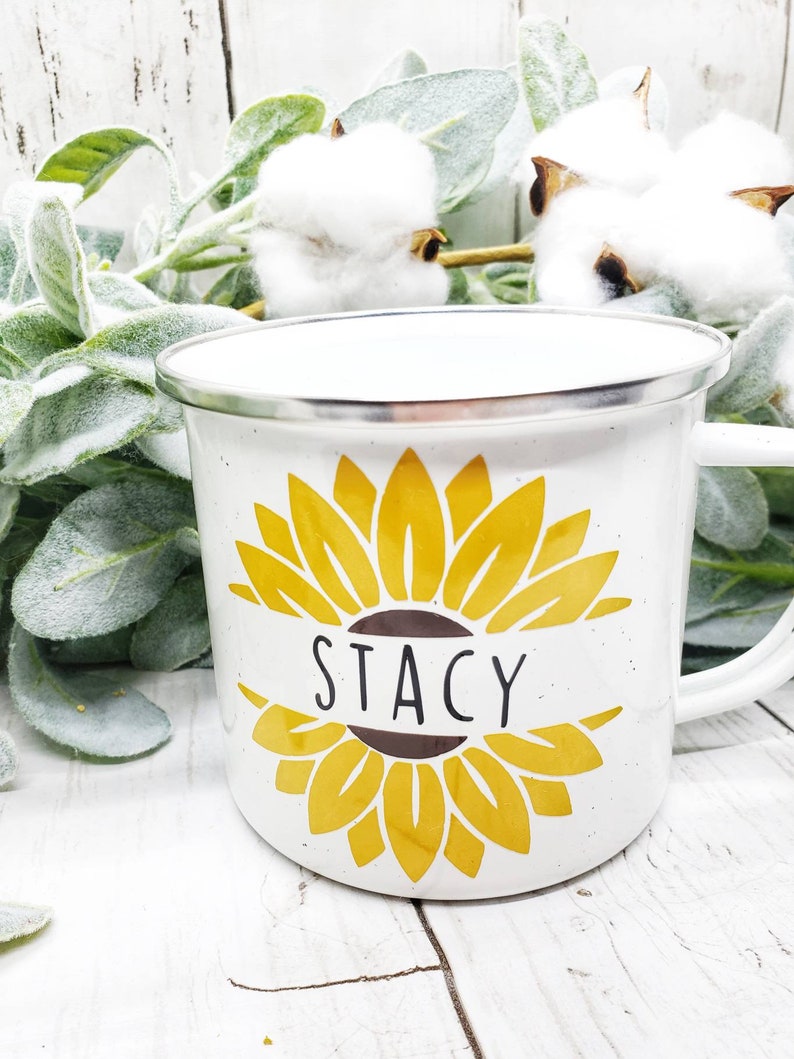 Op de afbeelding: Witte emaille mok met een geel zonnebloemontwerp en de naam "Stacy" op een bruin lint.