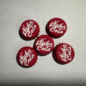 Könnte beinhalten: Fünf rote Kronkorken mit dem weißen Schriftzug "Nuka Cola". Die Kronkorken sind kreisförmig angeordnet und haben einen gewellten Rand. Der Hintergrund ist weiß.