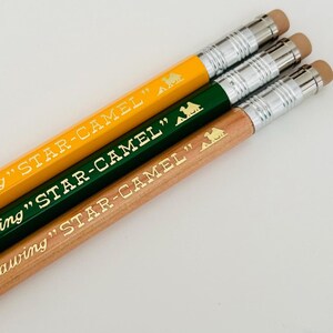 May include: Three mechanical pencils with different coloured barrels: yellow, green and natural wood. Each pencil has a gold camel logo and the text "STAR-CAMEL" printed on the barrel.