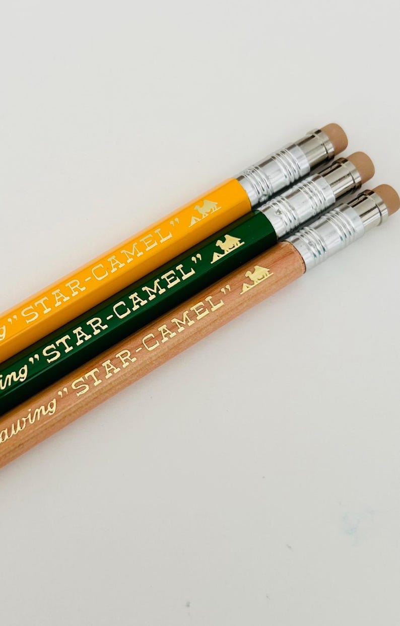 May include: Three mechanical pencils with different coloured barrels: yellow, green and natural wood. Each pencil has a gold camel logo and the text "STAR-CAMEL" printed on the barrel.