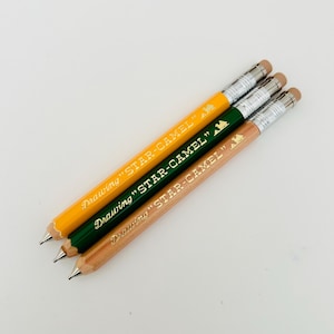 May include: Three mechanical pencils with wood, yellow, and green barrels. Each pencil has a gold inscription that reads "Drawing "STAR-CAMEL"".
