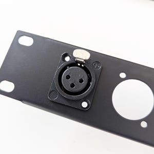 Puede incluir: Estante de metal negro con tres recortes circulares y un conector XLR de cinco pines.