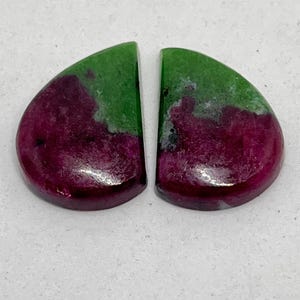 Może przedstawiać: Dwa polerowane kaboszony z kamieni szlachetnych, każdy o zaokrąglonym kształcie. Kamienie charakteryzują się żywą kombinacją kolorów głębokiej czerwieni i zieleni, z błyszczącym wykończeniem. Kamienie szlachetne są prawdopodobnie przeznaczone do tworzenia biżuterii lub kolekcjonowania.