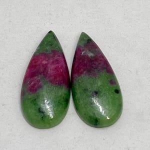 Może przedstawiać: Dwa kamienie szlachetne w kształcie łezki. Każdy kamień ma żywą mieszankę kolorów: głęboki magenta środek otoczony nakrapianym zielonym zewnętrzem. Kamienie są wypolerowane i gładkie, o naturalnym, organicznym wyglądzie. Kamienie szlachetne są na białym tle.