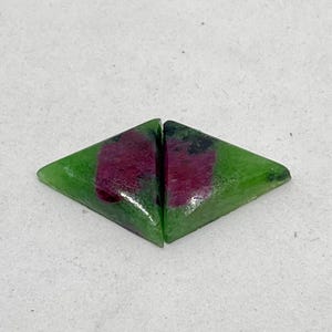 Puede incluir: Dos gemas verdes en forma de diamante con un centro rojo y morado. Las gemas están sobre un fondo blanco. Es probable que estas gemas se utilicen para la fabricación de joyas.