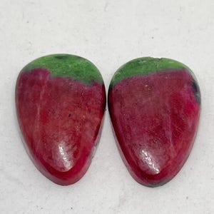 Könnte beinhalten: Zwei polierte, tropfenförmige Edelsteine. Der untere Teil jedes Steins ist tiefrot und geht in einen grünen oberen Bereich über. Die Steine werden vor einem weißen Hintergrund präsentiert.