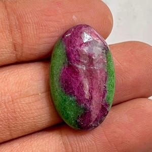 Puede incluir: Una gema de forma ovalada con una paleta de colores vibrantes. La piedra presenta un área central de magenta intenso, rodeada por una banda verde, creando un contraste llamativo. La superficie pulida refleja la luz, resaltando la belleza natural de la piedra.