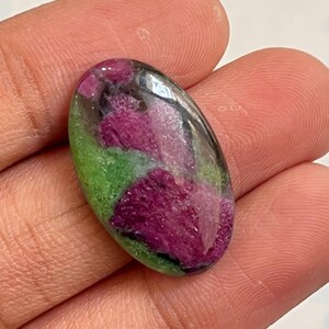 Puede incluir: Una gema ovalada con una superficie pulida. La piedra presenta una vibrante mezcla de colores, incluyendo magenta intenso, verde y negro. Las áreas magenta son prominentes, con acentos verdes y negros. La gema se sostiene en una mano.
