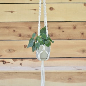 May include: A white macrame plant hanger with a green plant in a gray pot. The hanger is hanging from a wooden wall.