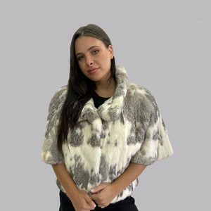 May include: A white and grey fur jacket with a short sleeve design. The jacket has a collar and a button closure.