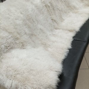 May include: A white sheepskin rug with a soft, fluffy texture. The rug is folded in half and is lying on a black leather surface.