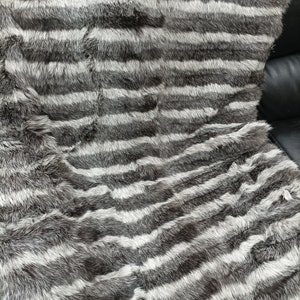 Puede incluir: Una manta de piel rayada con bandas alternas de piel gris oscuro y blanca. La piel es suave y esponjosa, y la manta es perfecta para añadir un toque de lujo a cualquier habitación.