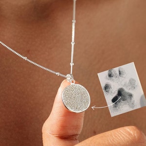 May include: A silver necklace with a round pendant featuring a paw print design. The pendant is made from a textured metal and is a unique way to remember a beloved pet.