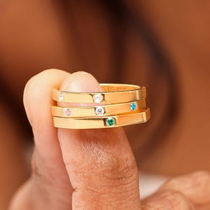 May include: A stack of four gold-colored rings, each with a thin band. The rings are adorned with small, colorful gemstones: a clear stone, a pink stone, a blue stone, and a green stone. The rings are stacked on a finger.
