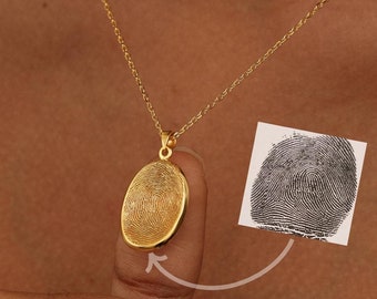 May include: A gold-colored oval pendant necklace with a fingerprint design. The pendant hangs from a delicate chain. A fingerprint is shown next to the pendant. The necklace is a personalized jewelry item.