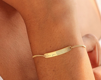 Puede incluir: Una pulsera dorada con una barra rectangular y una cadena delicada. La barra tiene una superficie texturizada y la inscripción "I love you". La pulsera se lleva en la muñeca, con una parte superior blanca en el fondo.