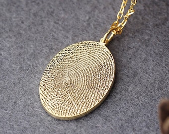 Könnte beinhalten: Goldfarbene ovale Anhänger Halskette mit detailliertem Fingerabdruck-Design. Der Anhänger hängt an einer zarten Goldkette. Die Halskette wird auf einer grauen Oberfläche mit Naturholz und Tannenzapfen im Hintergrund präsentiert.