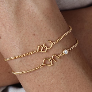 May include: Two gold chain bracelets with a small clear stone on one bracelet. The top bracelet has the letters "B" and "C" intertwined. The bottom bracelet has the letters "D" and "M" intertwined.