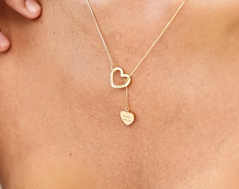 May include: A delicate gold necklace featuring two heart pendants. The upper heart is an open design, while the lower heart pendant has engraved text. The necklace is worn on a person's neck.