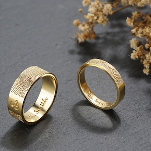 A couple gold ring is standing on a black stone. One ring for men, which is 7mm wide, and one ring for woman, which is 4mm wide. The fingerprint is engraved outside of the ring, and the inscription is engraved inside of the ring.