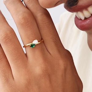 May include: A gold ring featuring a small, heart-shaped green gemstone and a white gemstone. The ring is on a person's finger, with the person's hand and face visible in the image.